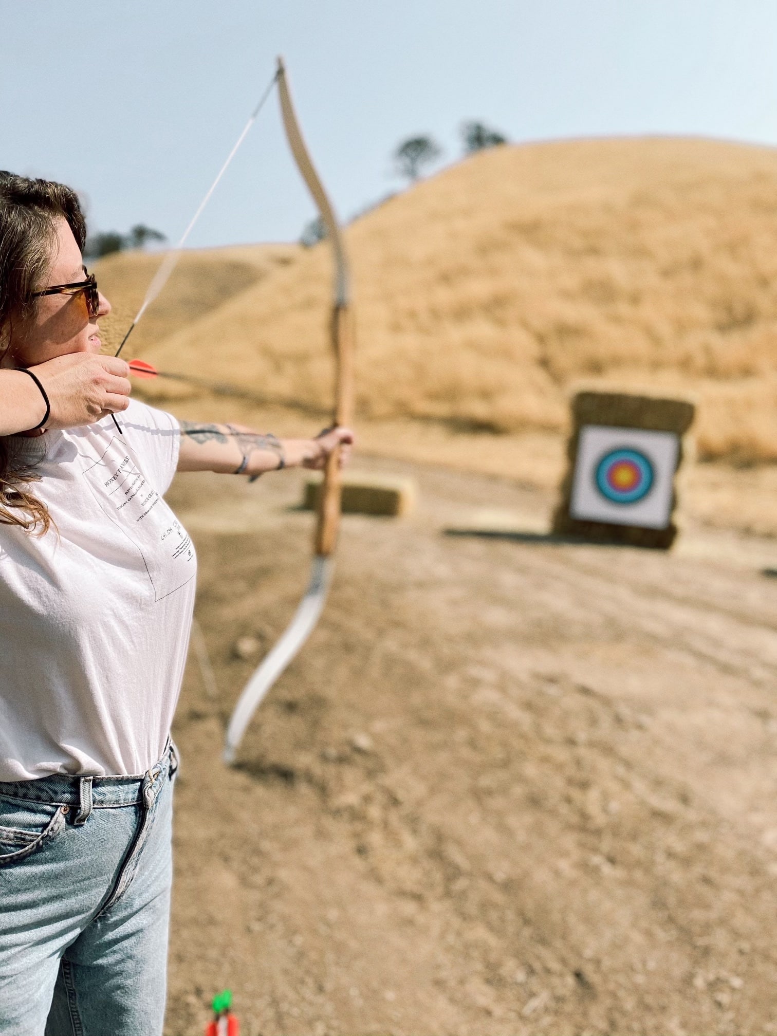 A focused woman practicing archery at Cass Winery during the Camp Cass program, with a target in the distance on a sunny day.