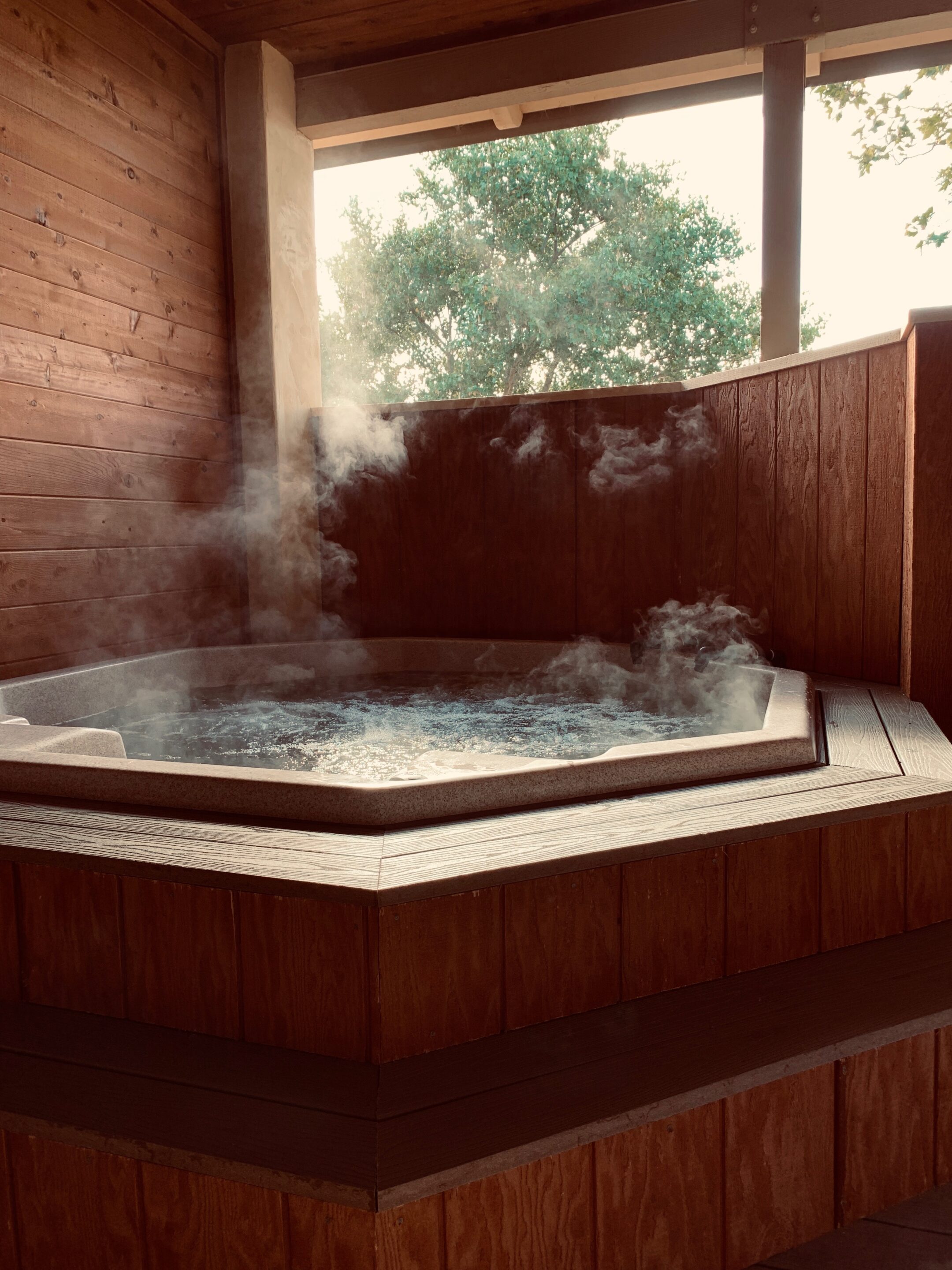 A steaming hot spring tub encased in a wooden structure with a view of leafy green trees through an open window, suggesting a serene spa experience.